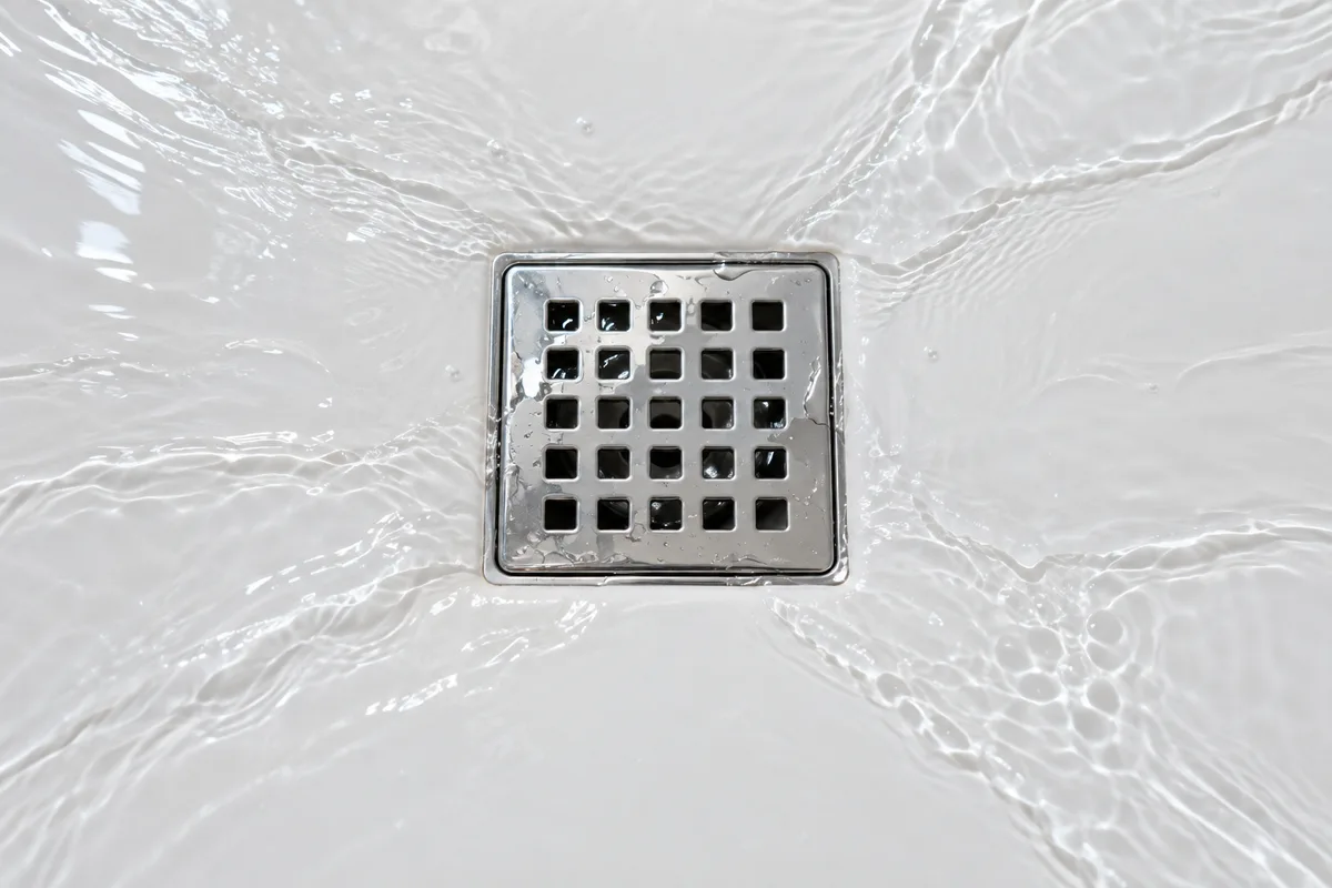 Grille d&rsquo;évacuation chromée carrée au centre d&rsquo;un receveur de douche blanc, eau s&rsquo;écoulant en nappe